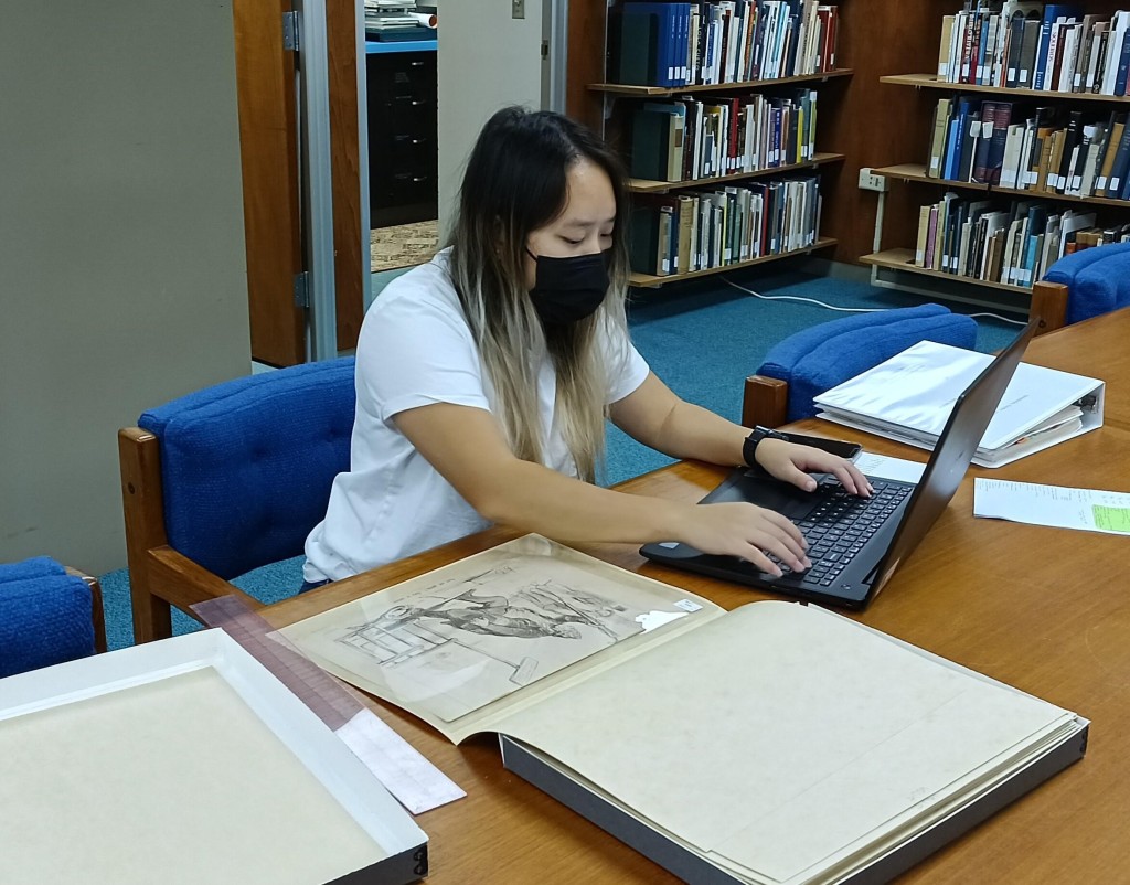 Student assistant Seng Khang seated at the table in the Jean Charlot Collection Reading Room, entering data about the print she is examining into a laptop computer.